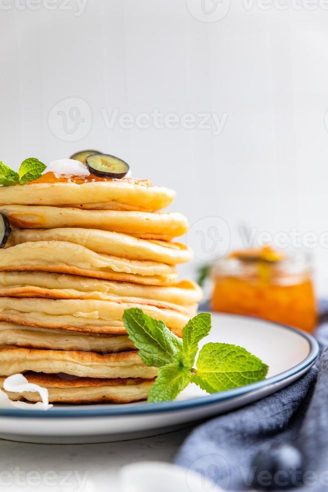 pila de panqueques esponjosos con mermelada de naranja, arándanos, chips de coco y menta, fondo claro. desayuno tradicional. fotografía de alta clave. foto