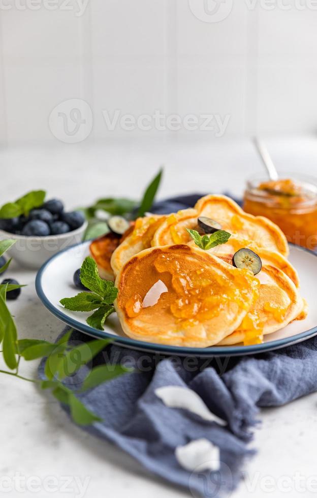 Traditional fluffy pancakes with orange jam, blueberries, coconut chips and mint for healthy breakfast, light background. High key photography. photo