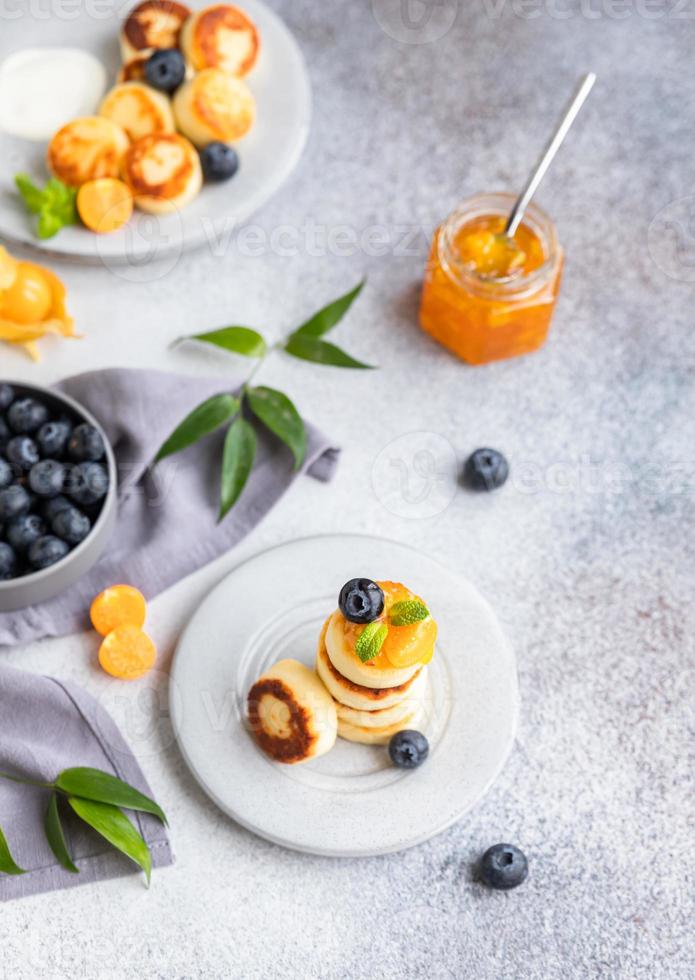 tortitas de requesón, buñuelos de cuajada dulce con arándanos y physalis, fondo de hormigón. syrniki con mermelada y bayas para un desayuno saludable. foto