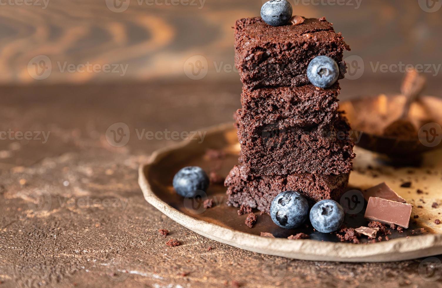 Chocolate brownie squares decorated with blueberry and pieces of dark chocolate and cocoa powder on craft ceramic plate, brown background. photo