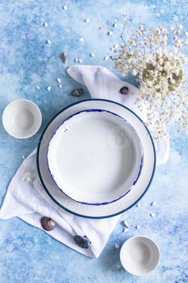 Handmade ceramic white plates with blue stripe on the edge, dried flowers and empty seashells on blue concrete background. photo