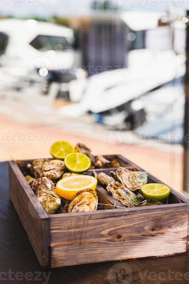 A variety of fresh oysters with lime and lemon in a wooden box. Fresh seafood. Outdoor cafe terrace. Blurred background with view of the yacht club. photo