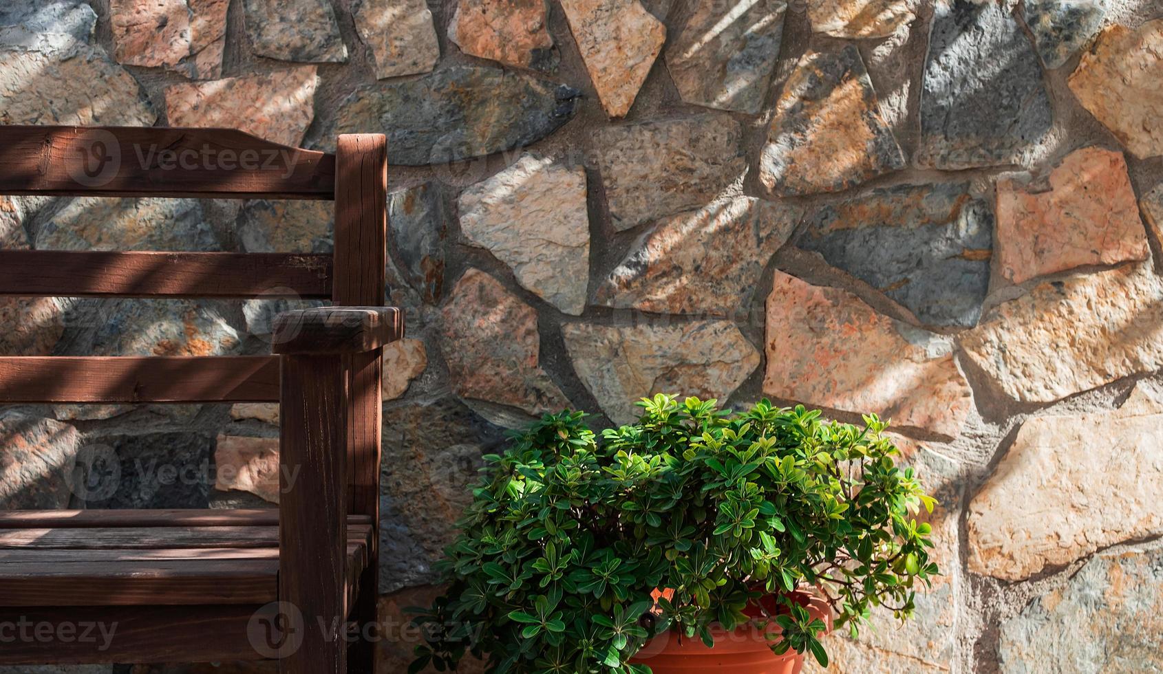 Fragment of a wooden bench against the background of a stone wall and a bush in a pot, the details of the design of the space of the yard or patio, shadows from tree crowns on the wall photo