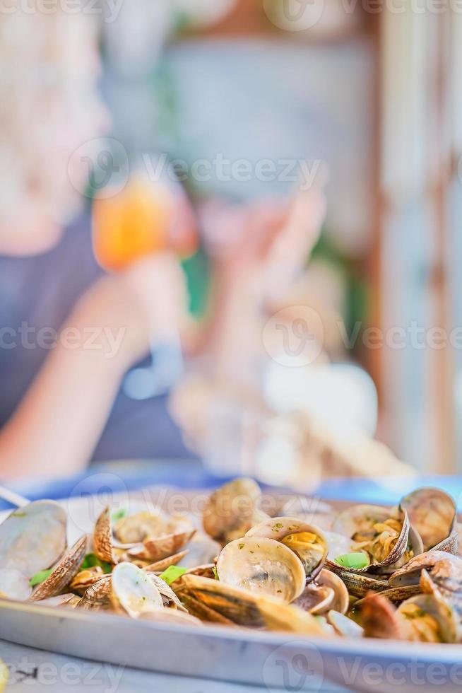 Blurred background with a dish of boiled vongole clams on a table in a cafe, vertical frame, in the background the silhouette of a woman with a glass of wine. Idea for a screensaver or banner photo