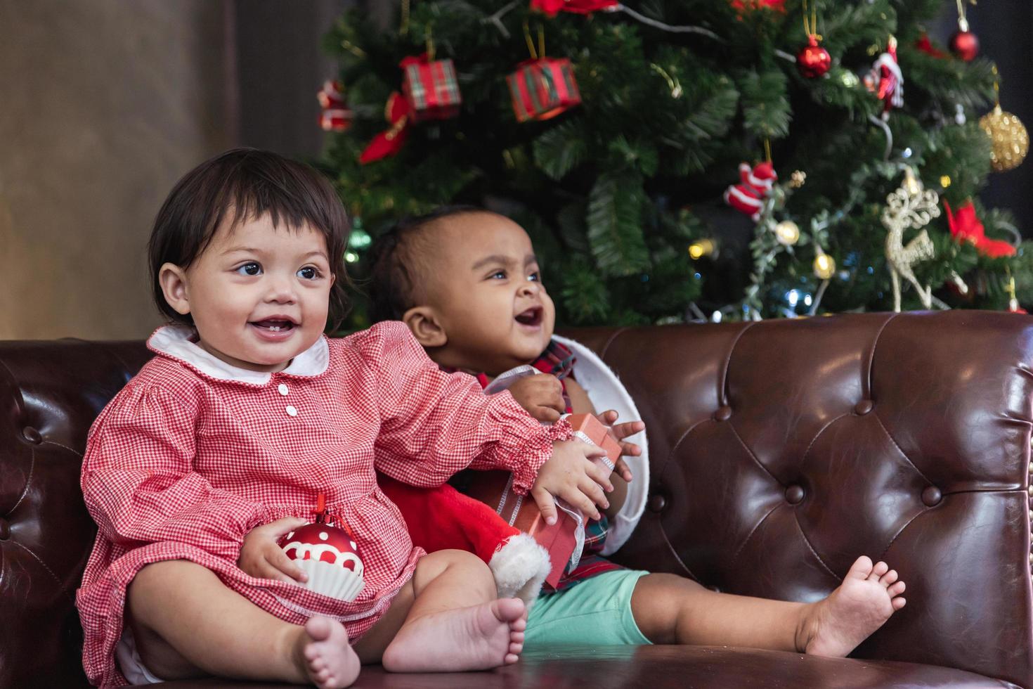 Two diverse baby friends are happily smiling while playing with present on couch dressing in christmas costume and santa hat with christmas tree on the back for season celebration concept photo