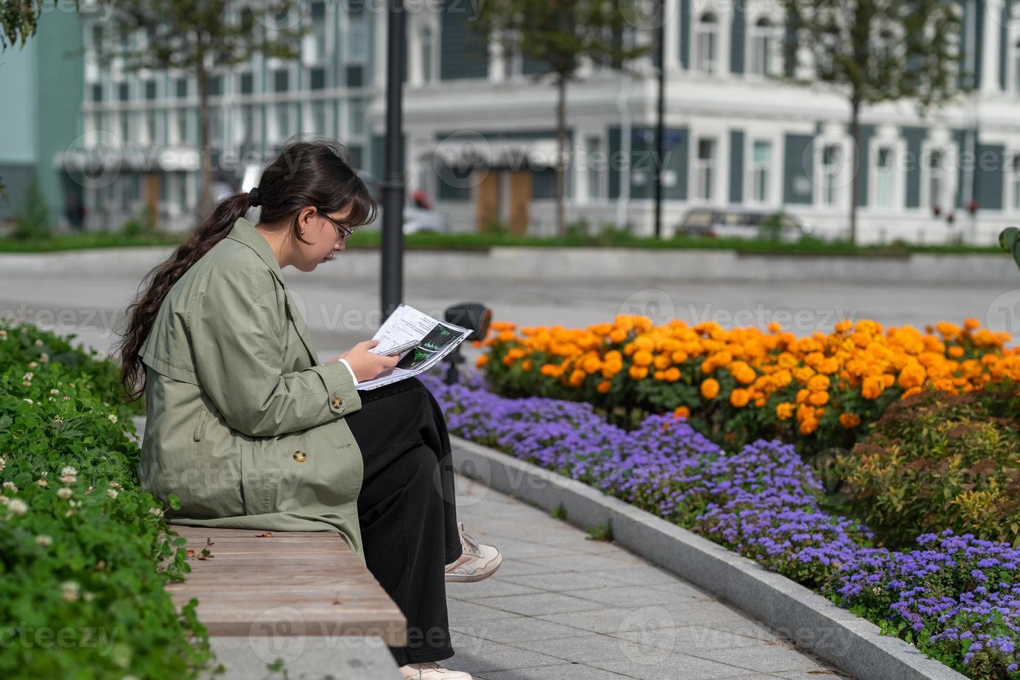 A girl sits on a bench on the street with documents in her hands and using her phone photo