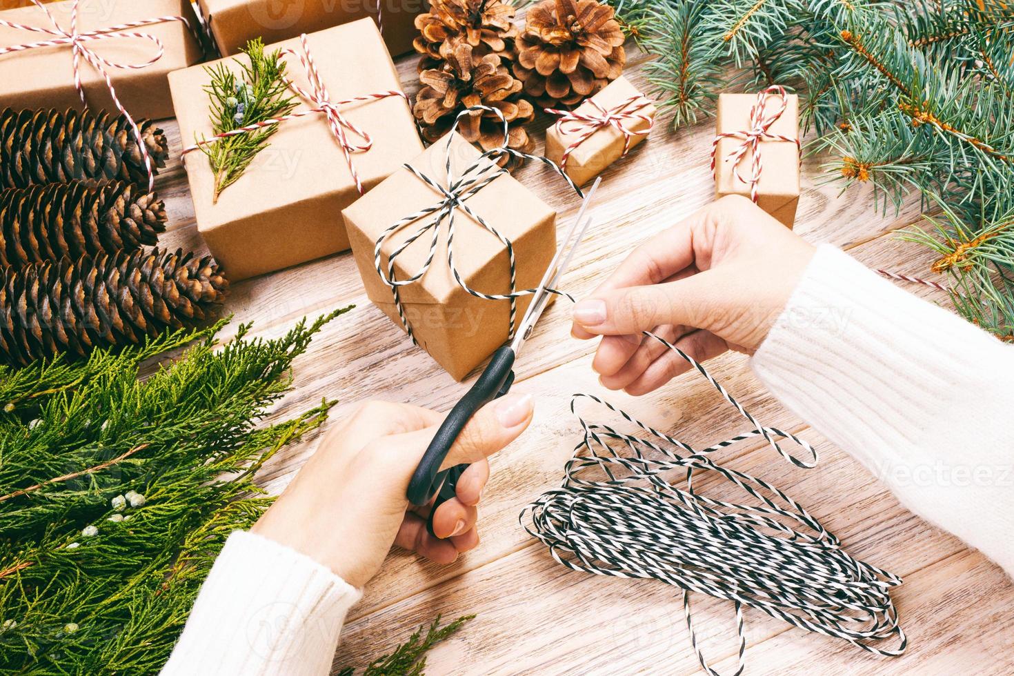 Hand crafted gift on wooden background with Christmas decor. Woman wrapping christmas present , girl prepares xmas gifts with fir tree and pine cone. Top view, copy space. Toned photo