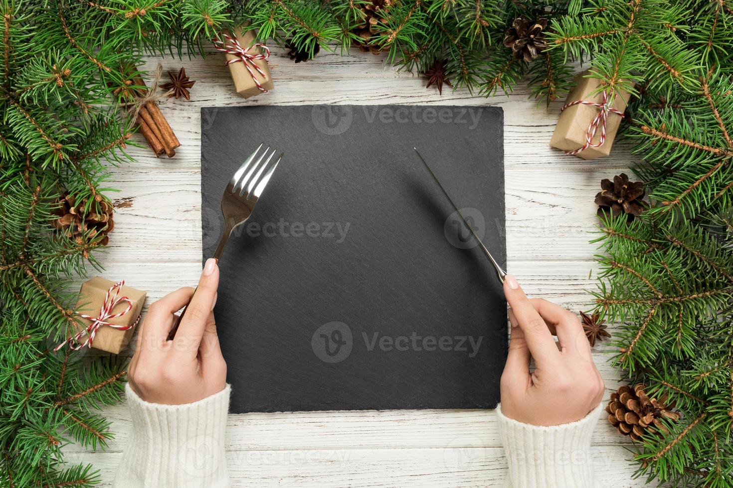 top view girl holds fork and knife in hand and is ready to eat. Empty black slate square plate on wooden christmas background. holiday dinner dish concept with new year decor photo