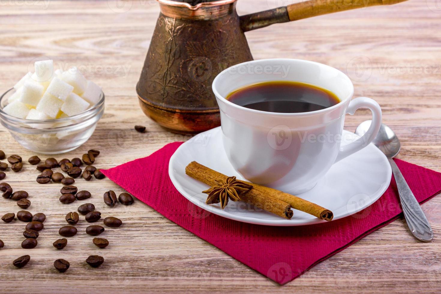 taza blanca de café negro caliente en el platillo, servida con granos de café, terrones de azúcar blanco en un bol, cuchara de té, anís y palitos de canela sobre fondo texturizado de madera foto