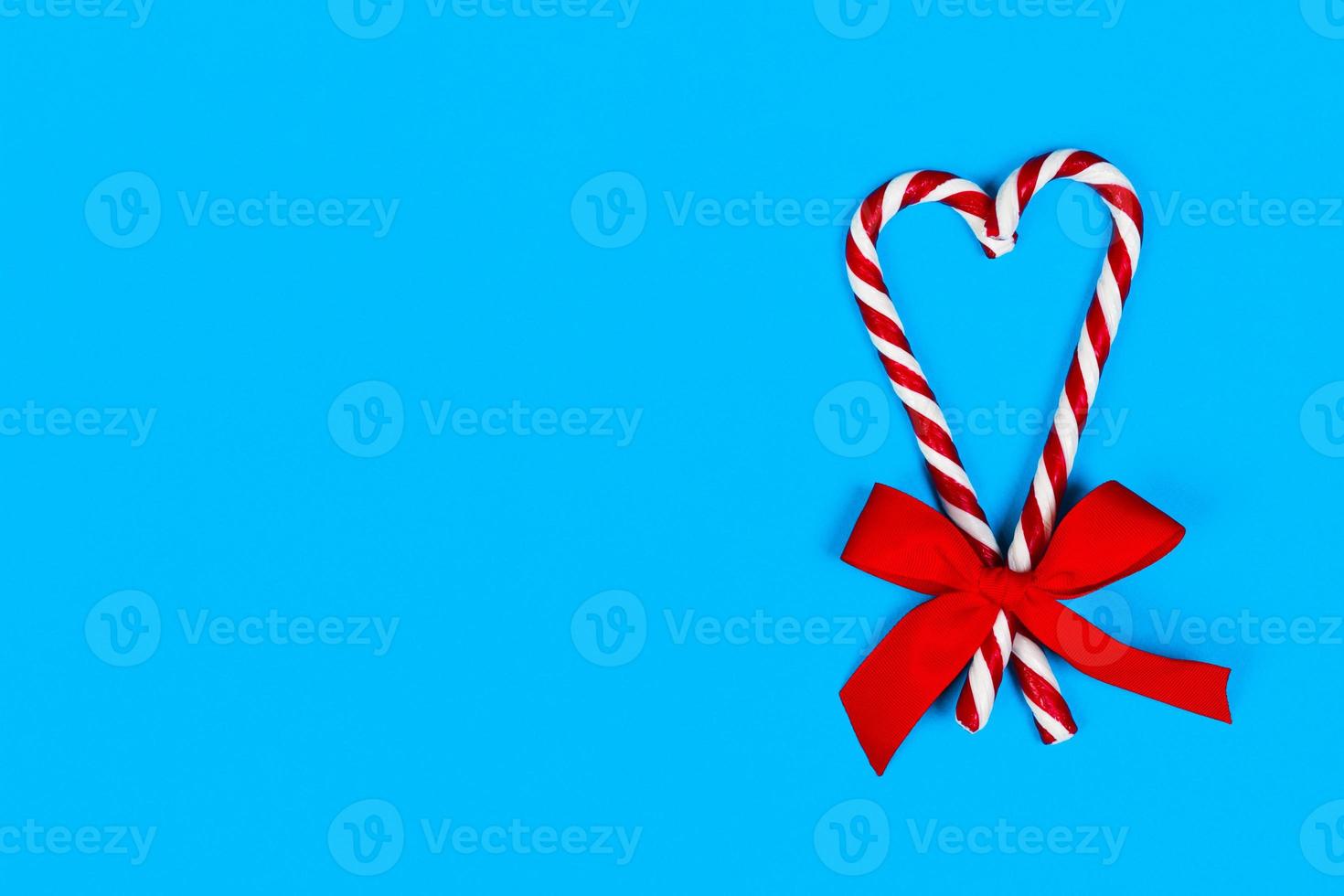 vista superior bastón de caramelo rayado en colores navideños en forma de corazón con lazo rojo sobre fondo azul. de cerca. Navidad, temporada de vacaciones. copie el espacio foto