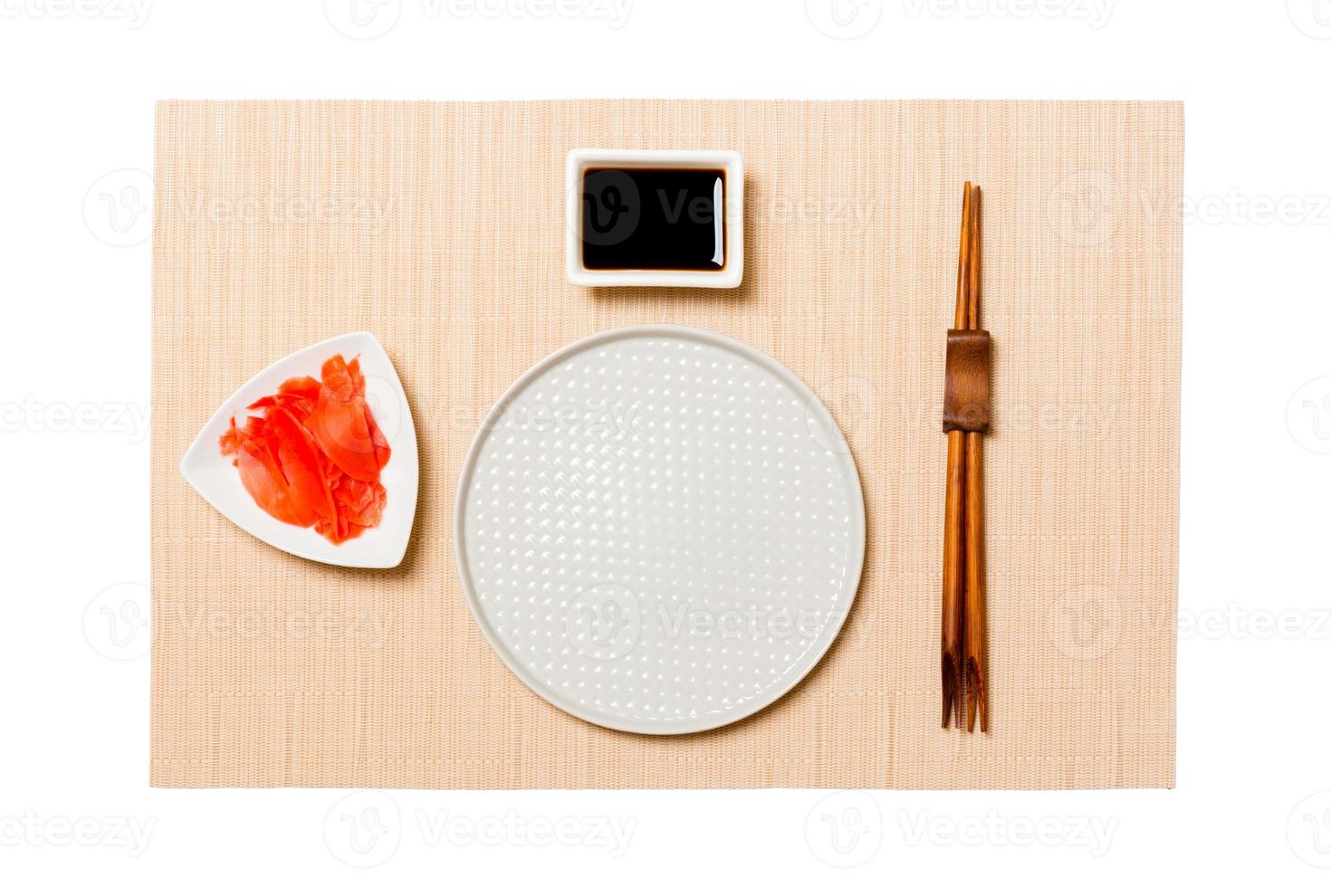 Empty round white plate with chopsticks for sushi, ginger and soy sauce on brown sushi mat background. Top view with copy space for you design photo