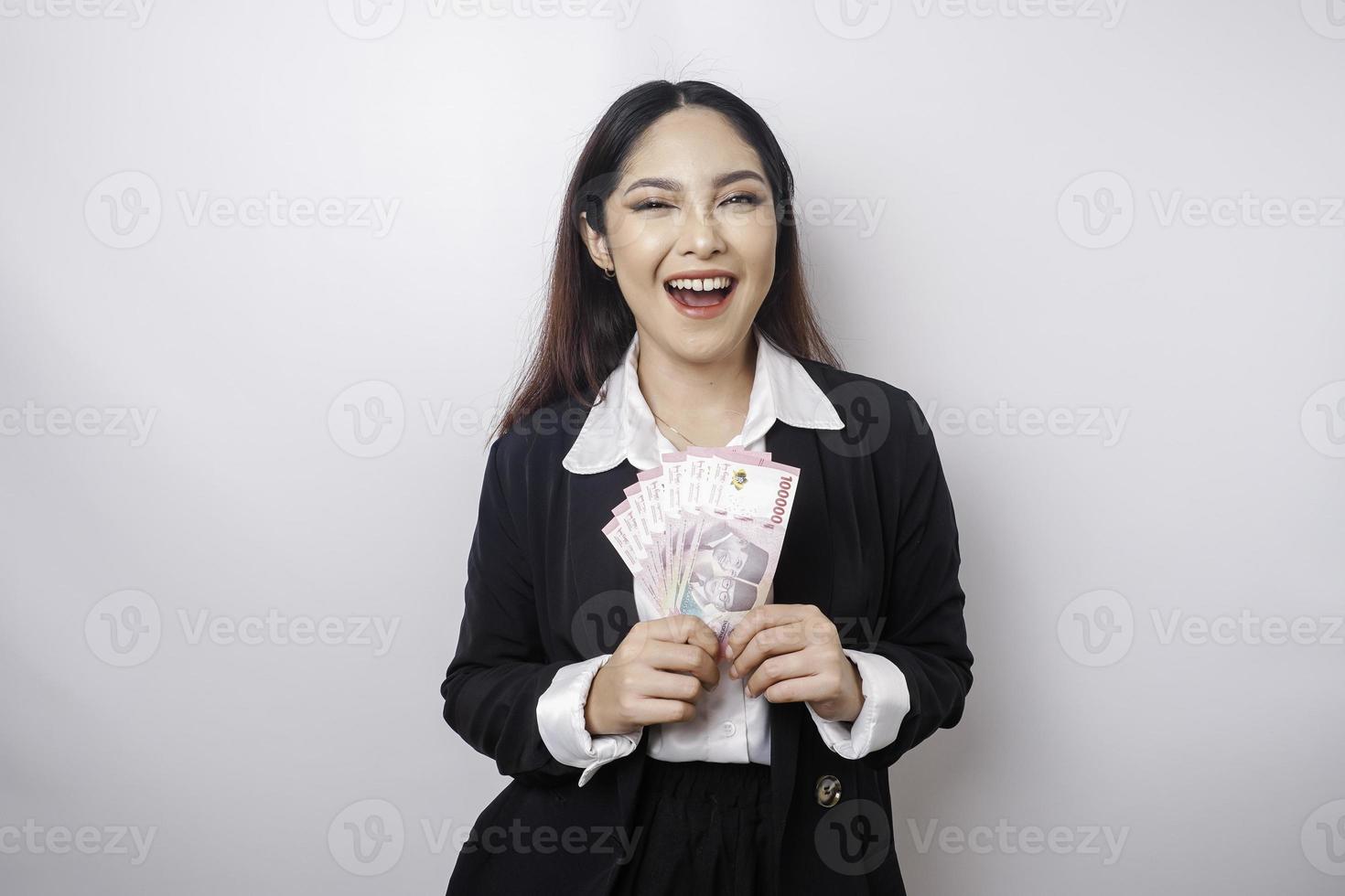 A happy young businesswoman is wearing black suit and holding cash money in Indonesian rupiah isolated by white background photo