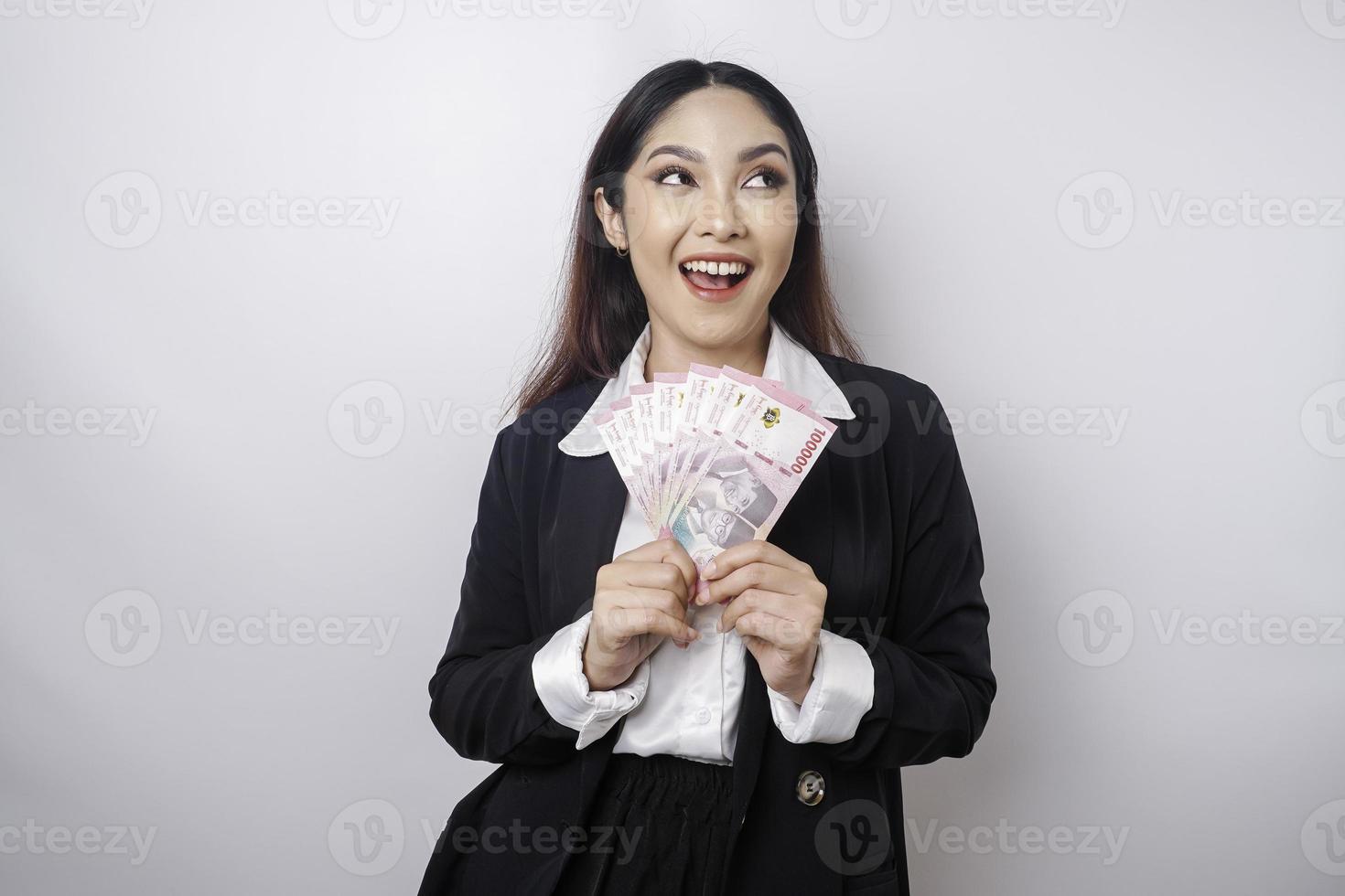 A thoughtful young woman is wearing black suit and holding cash money in Indonesian rupiah isolated by white background photo