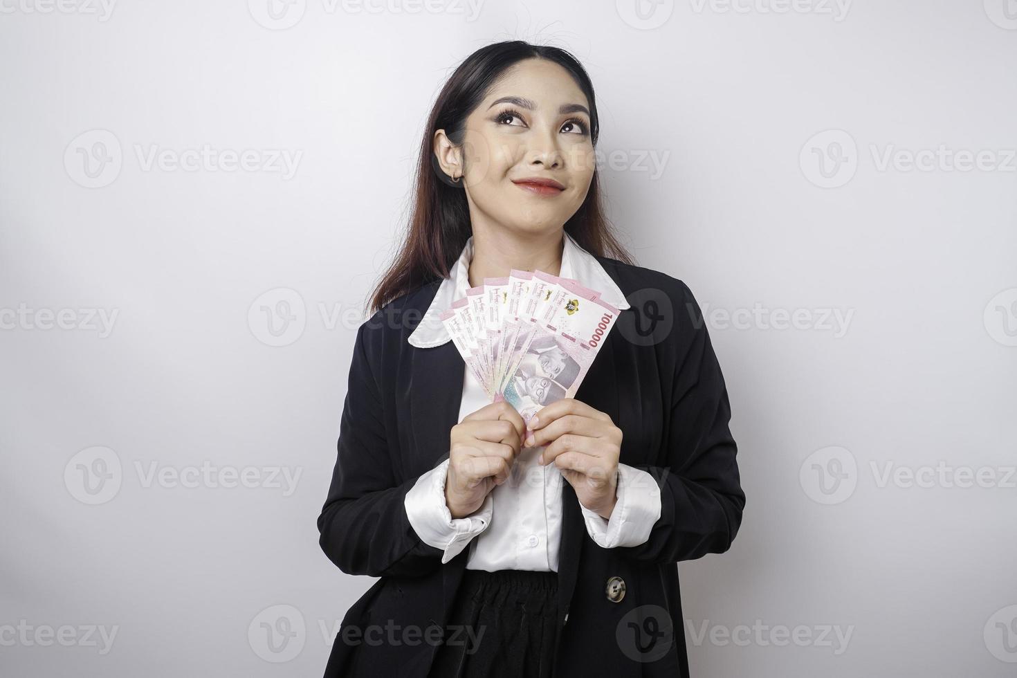 A thoughtful young woman is wearing black suit and holding cash money in Indonesian rupiah isolated by white background photo