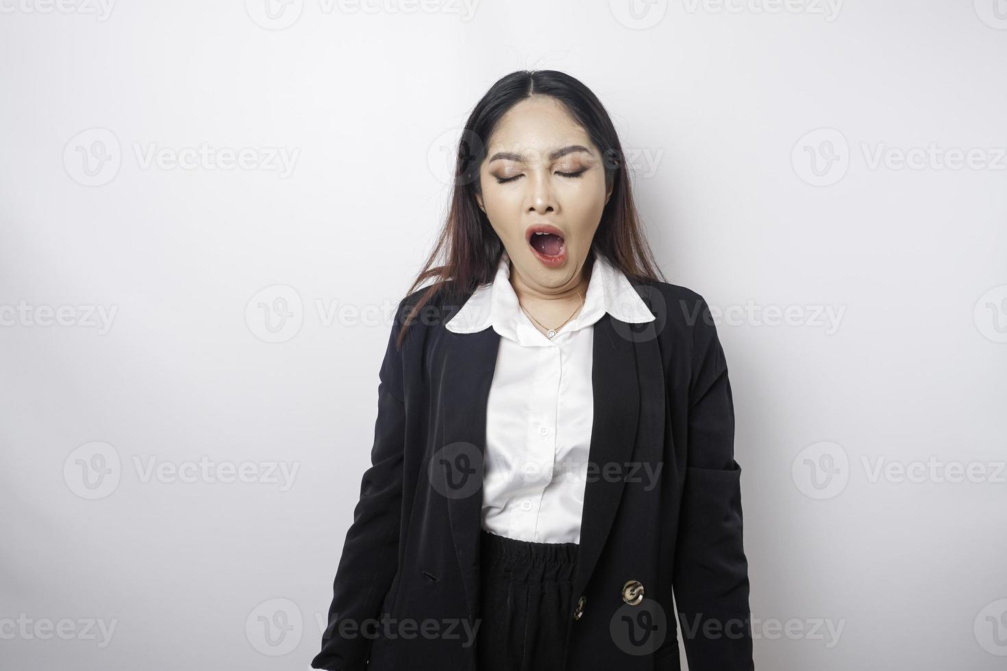 Portrait of sleepy attractive Asian woman wearing a black suit, feeling tired after overtime at work, yawning, covering opened mouth with palm photo