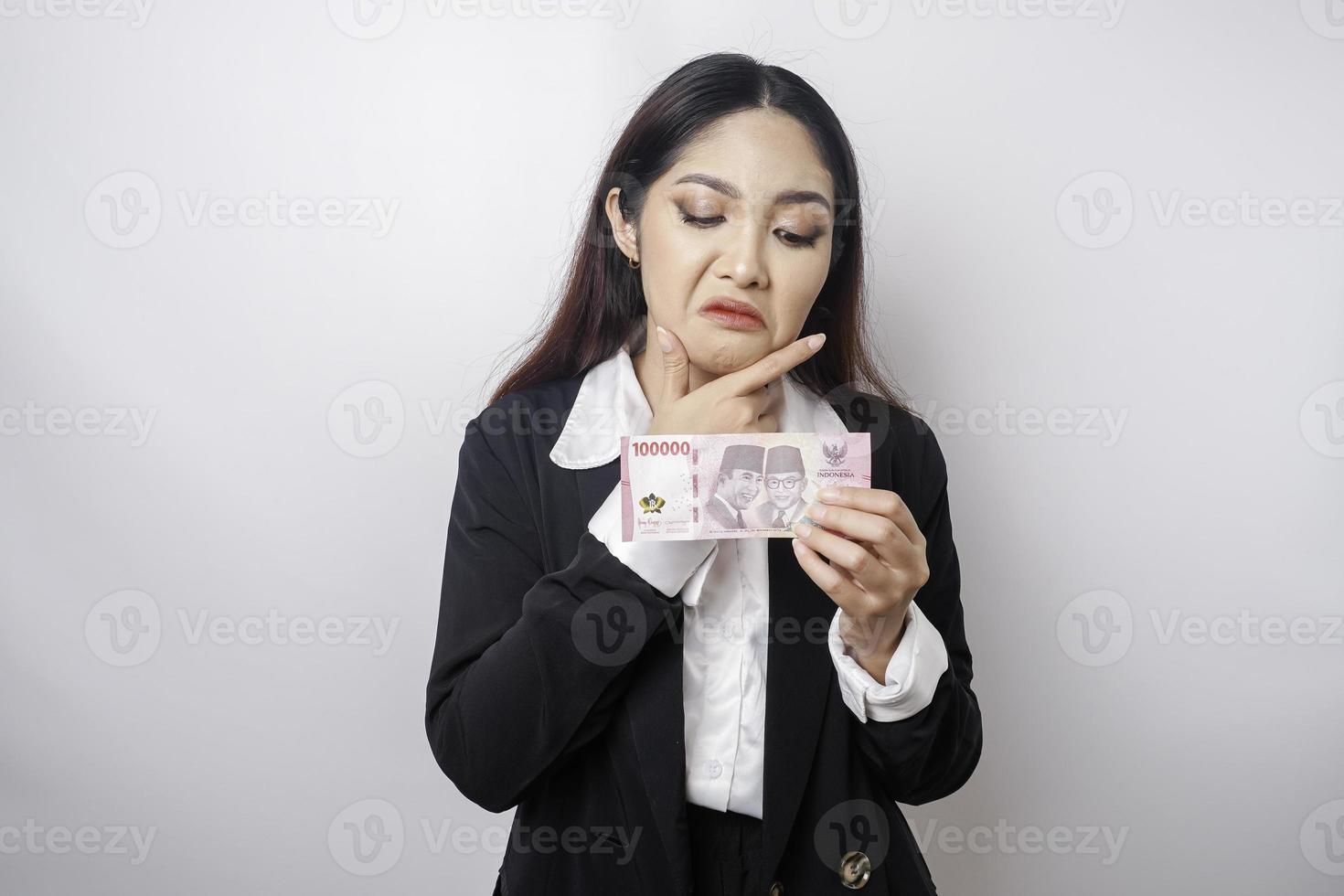 A thoughtful young woman is wearing black suit and holding cash money in Indonesian rupiah isolated by white background photo
