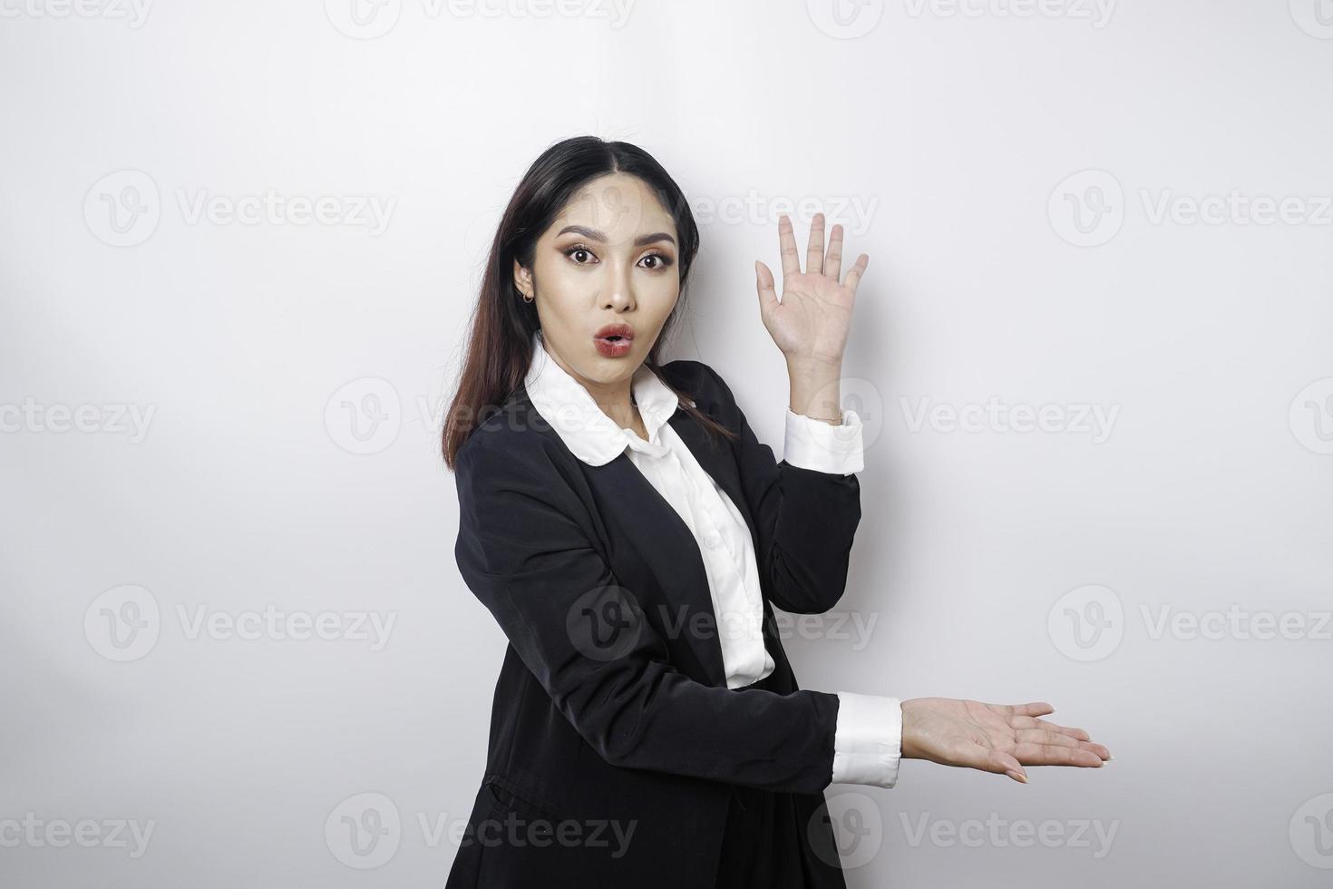 Shocked Asian businesswoman wearing a black suit pointing at the copy space beside her, isolated by a white background photo