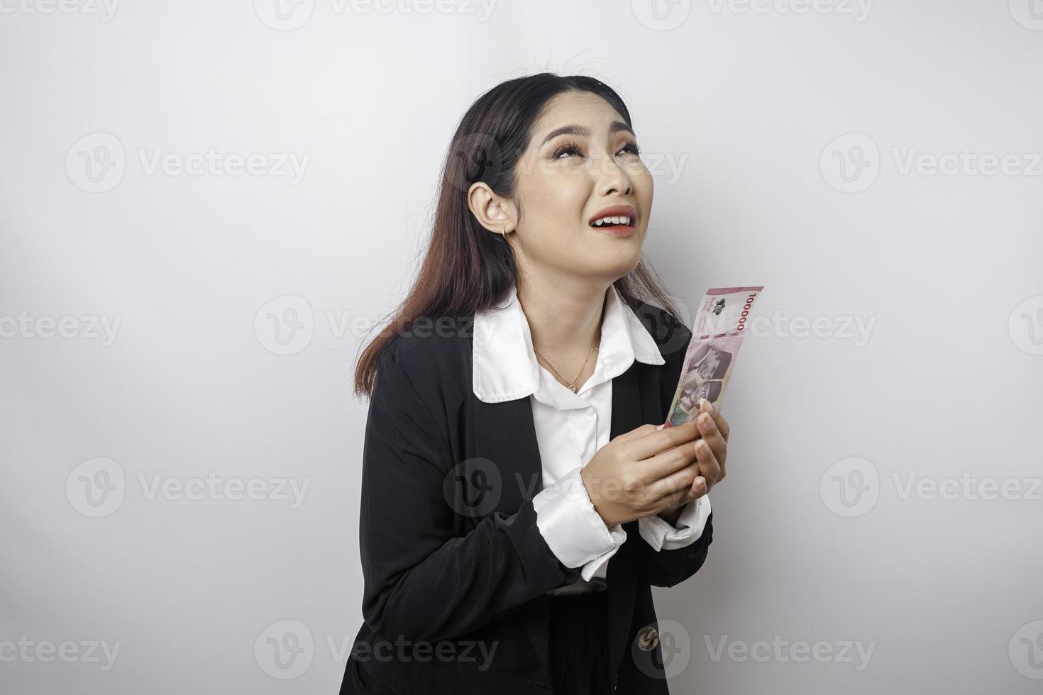 A portrait of an Asian businesswoman wearing a black suit and holding money in Indonesian Rupiah isolated by white background looks depressed photo