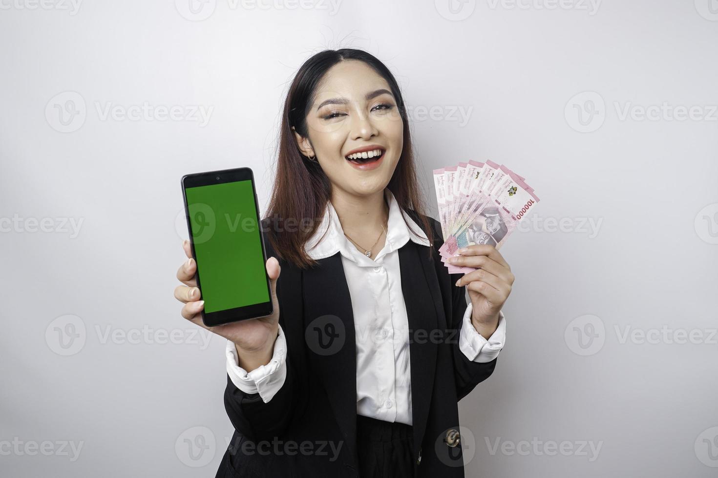 A happy young businesswoman is wearing black suit, showing her phone and money in Indonesian rupiah isolated by white background photo