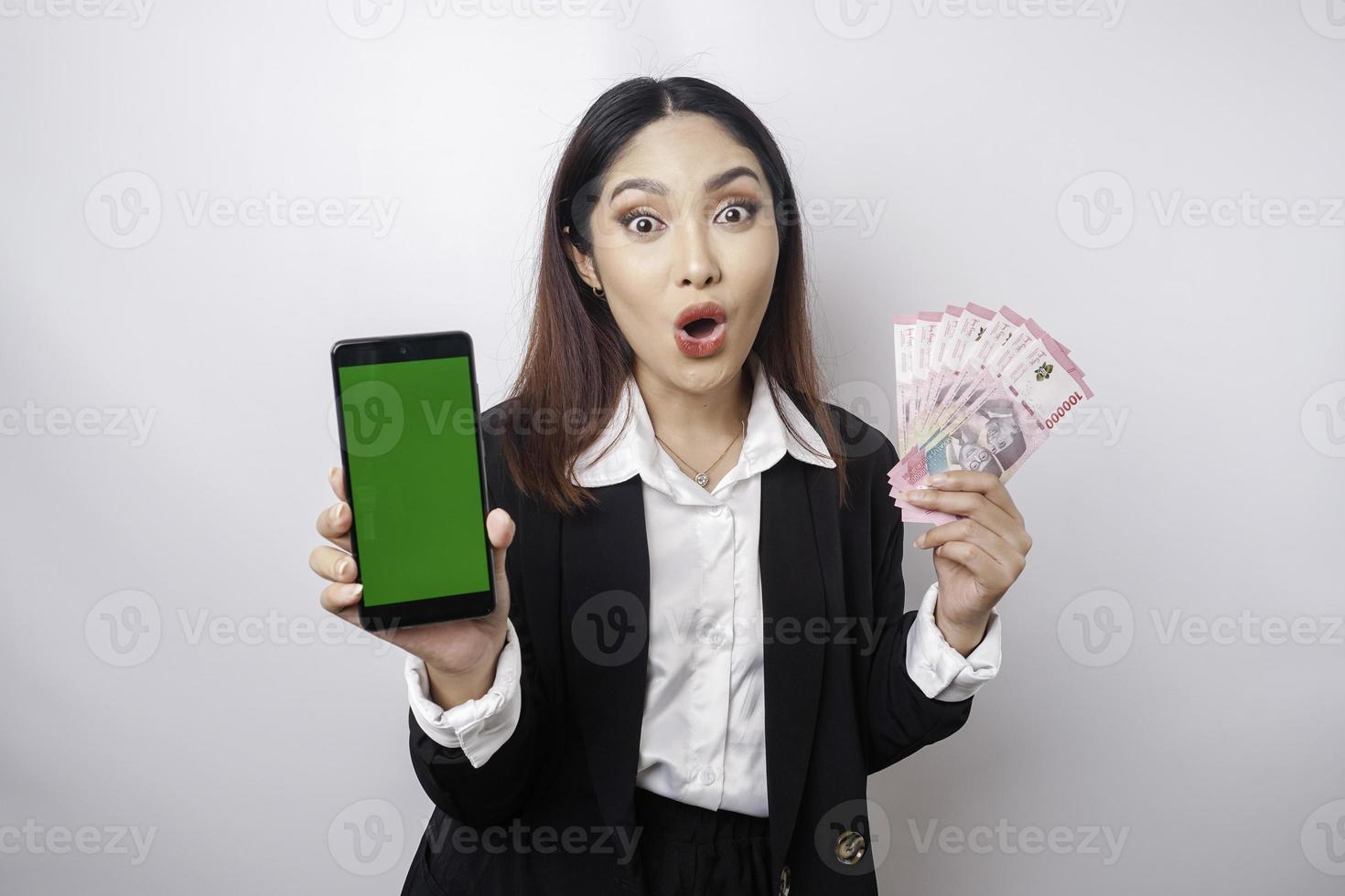 Surprised Asian businesswoman wearing black suit showing her smartphone and money in Indonesian Rupiah, isolated by white background photo