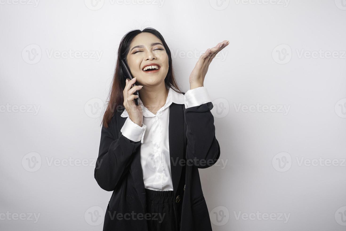 Excited Asian businesswoman wearing black suit pointing at the copy space on top of her while talking on a phone call, isolated by white background photo