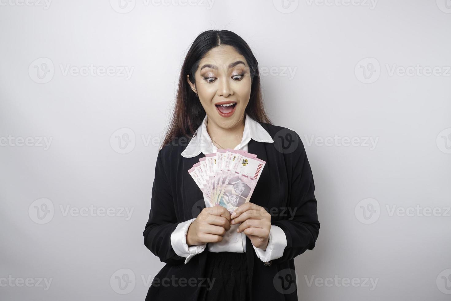 A happy young businesswoman is wearing black suit and holding cash money in Indonesian rupiah isolated by white background photo