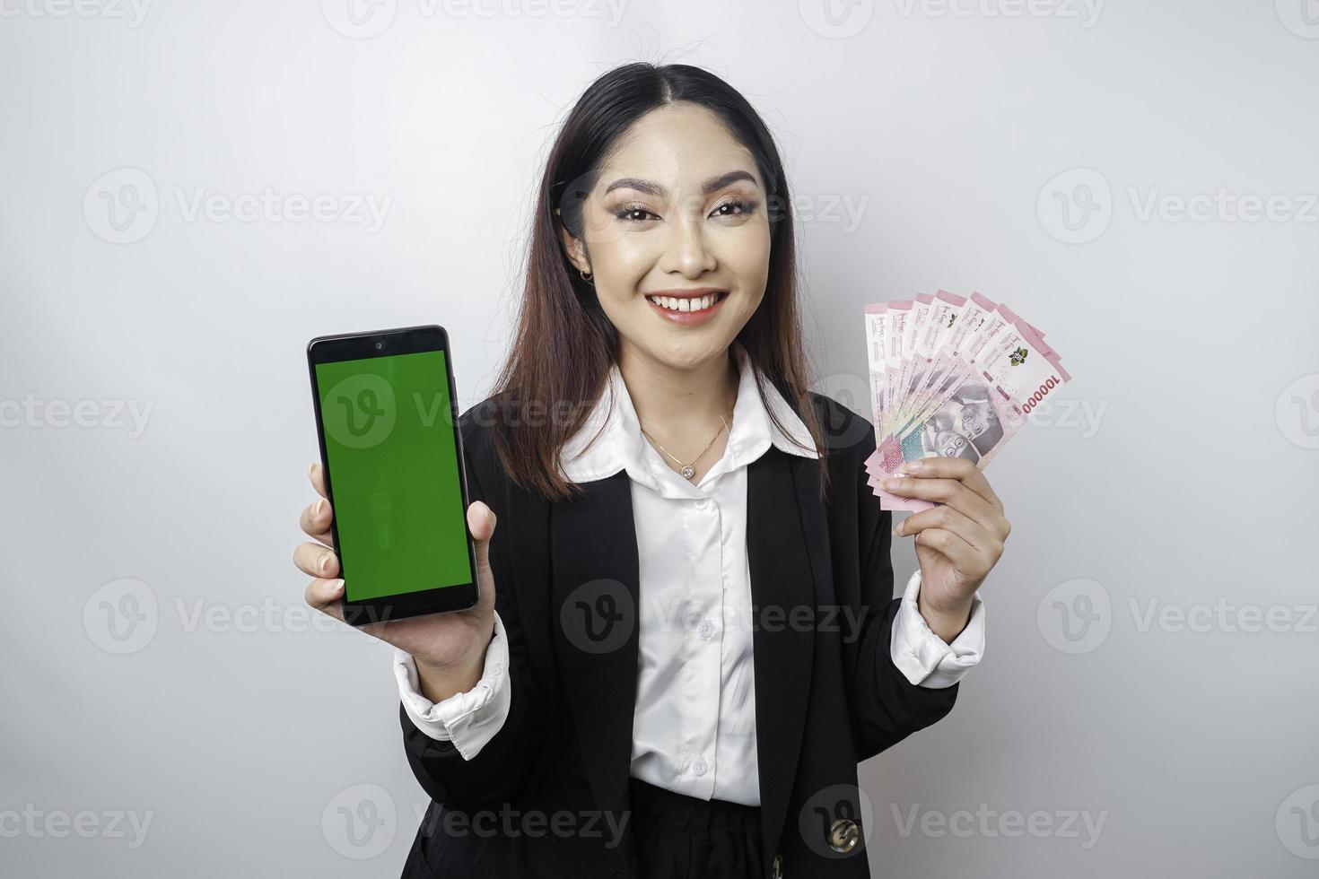 A happy young businesswoman is wearing black suit, showing her phone and money in Indonesian rupiah isolated by white background photo