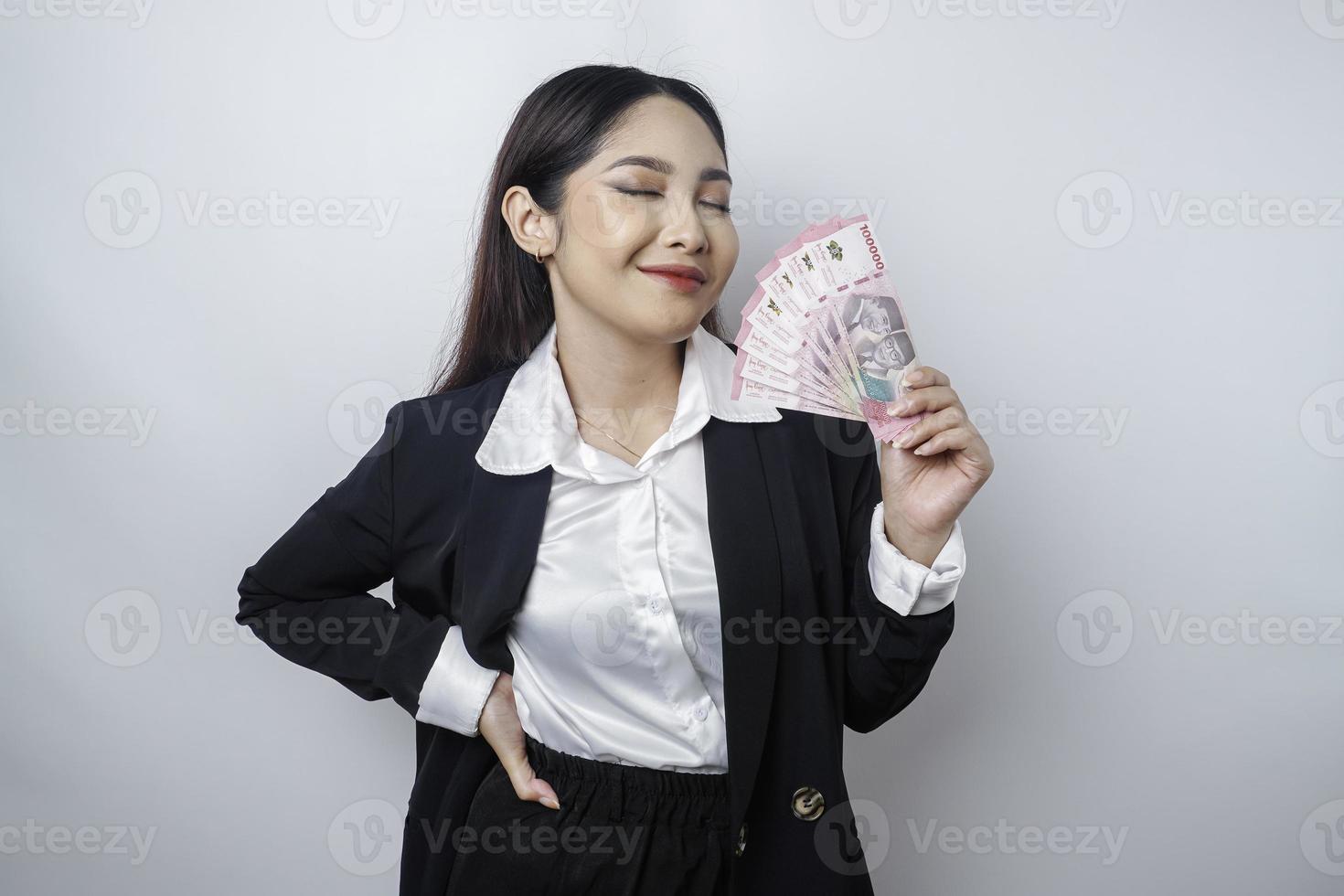 una joven empresaria feliz lleva traje negro y tiene dinero en efectivo en rupias indonesias aislada de fondo blanco foto