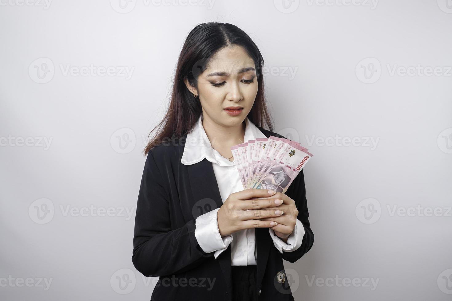 A confused young businesswoman is wearing black suit and holding cash money in Indonesian rupiah isolated by white background photo