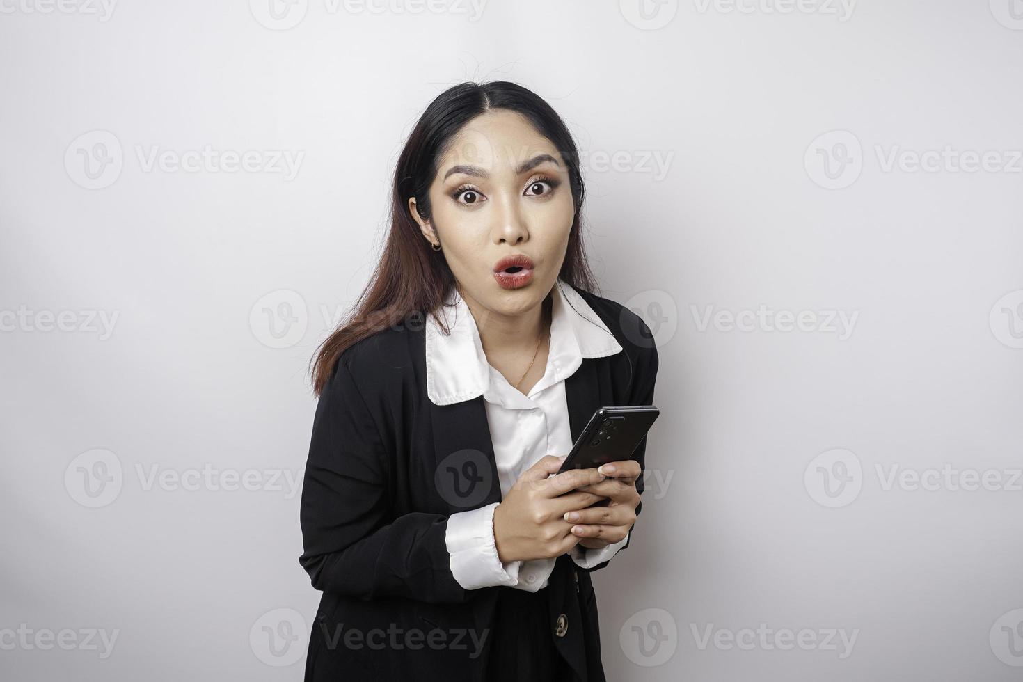 Surprised Asian businesswoman wearing black suit holding her smartphone, isolated by white background photo