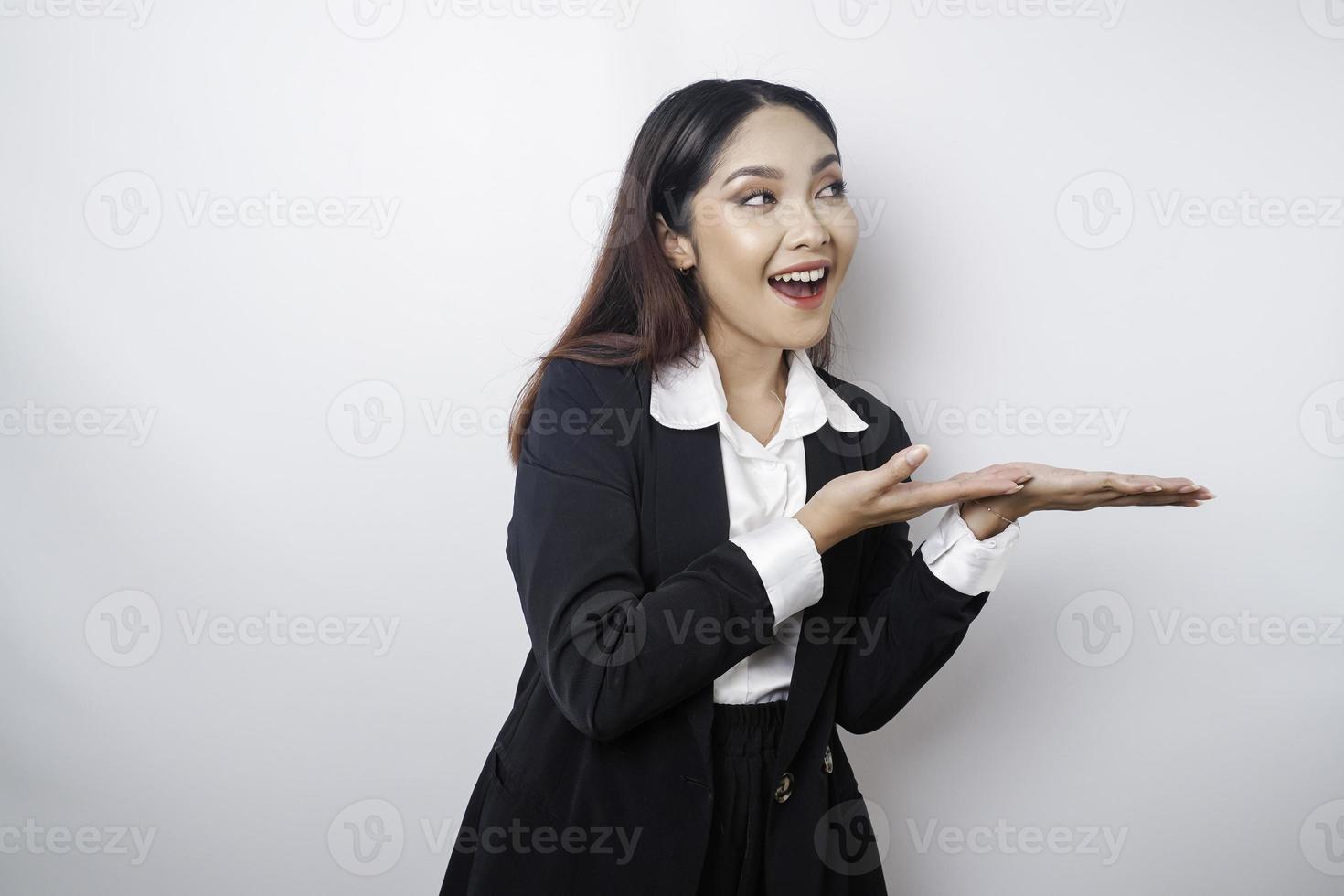 Excited Asian businesswoman wearing black suit pointing at the copy space beside her, isolated by white background photo