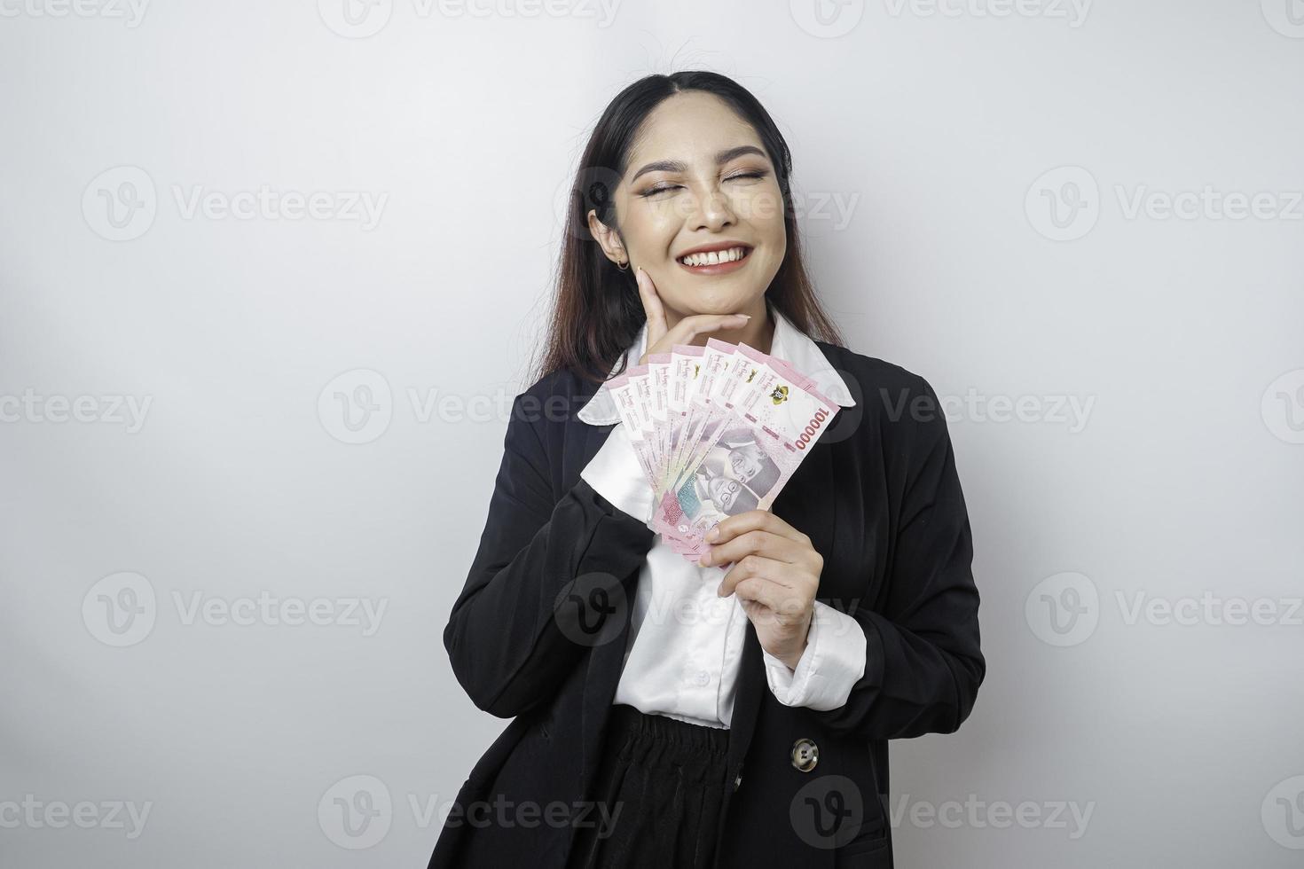 A thoughtful young woman is wearing black suit and holding cash money in Indonesian rupiah isolated by white background photo
