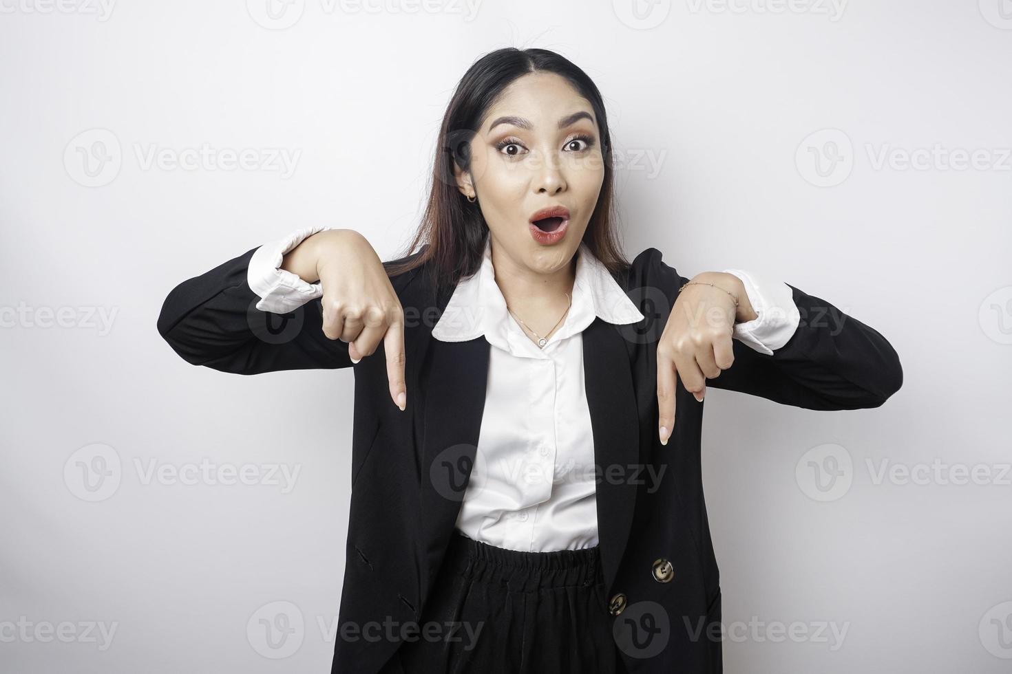 Shocked Asian businesswoman wearing a black suit pointing at the copy space below her, isolated by a white background photo