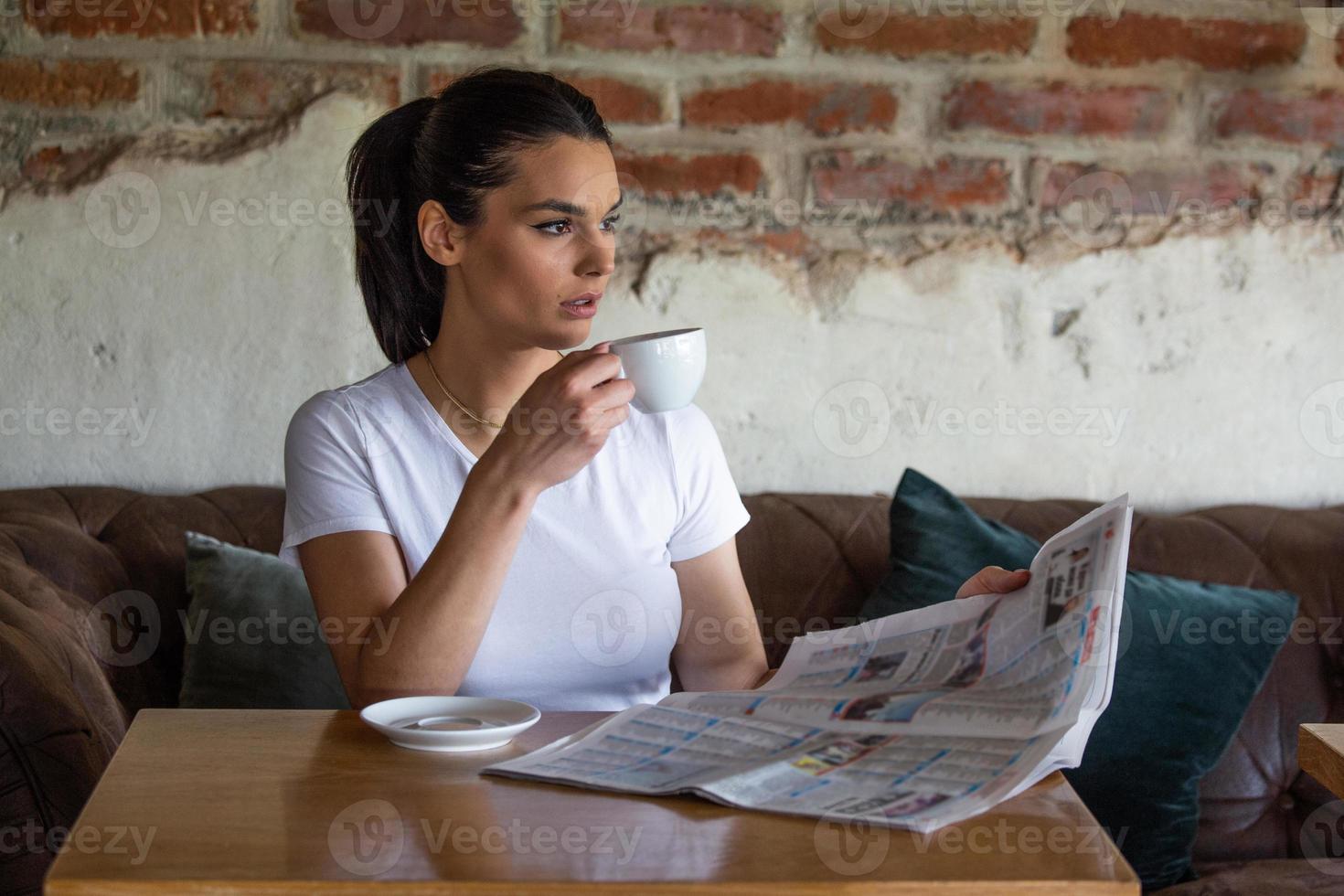Charming woman with beautiful smile reading newspaper during rest in coffee shop, happy Caucasian female reading news while relaxing in cafe during free time photo