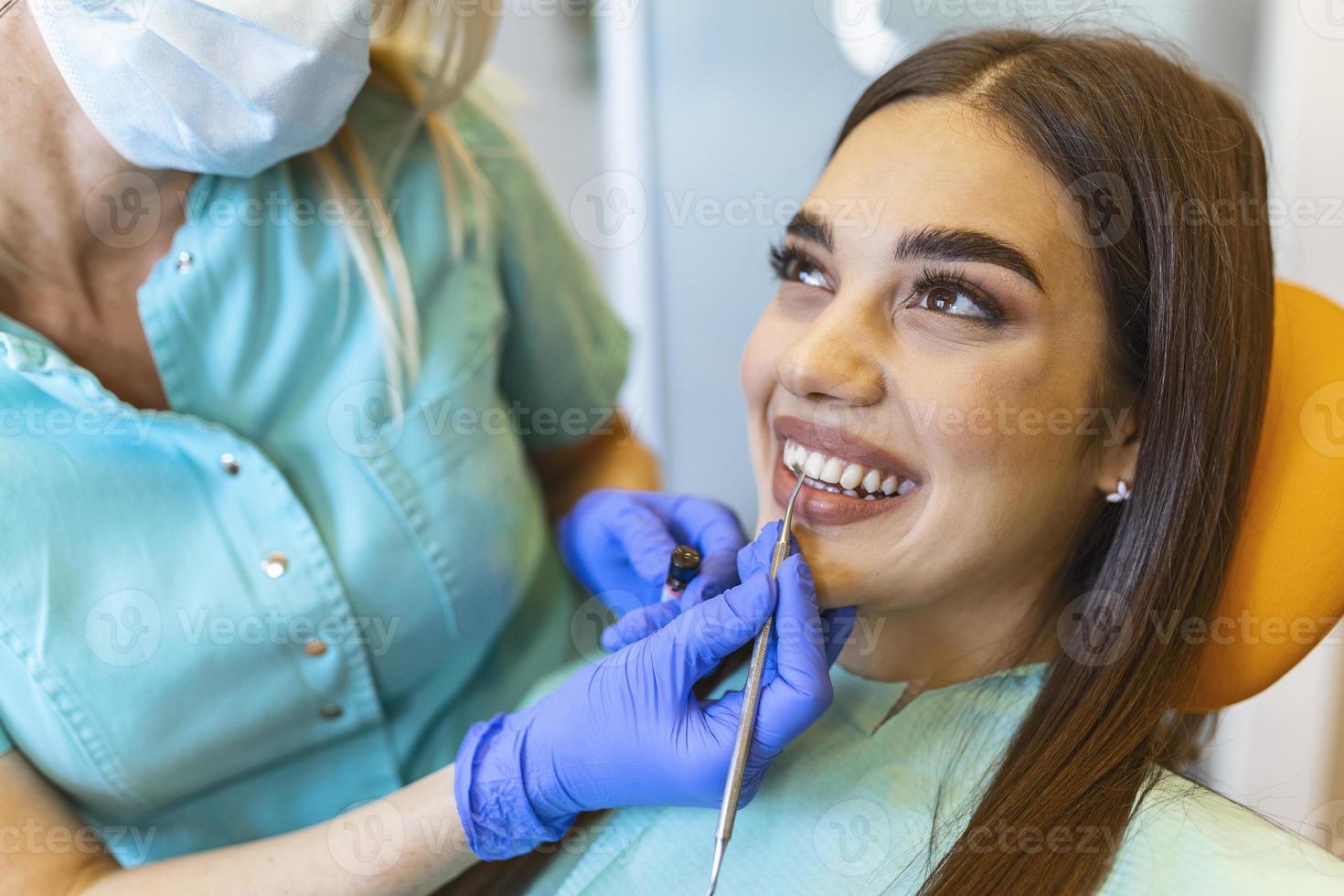 Image of satisfied young woman sitting in dental chair at medical center while professional doctor fixing her teeth, Female dentist choosing filling shade for smiling woman, using tooth scale sample photo