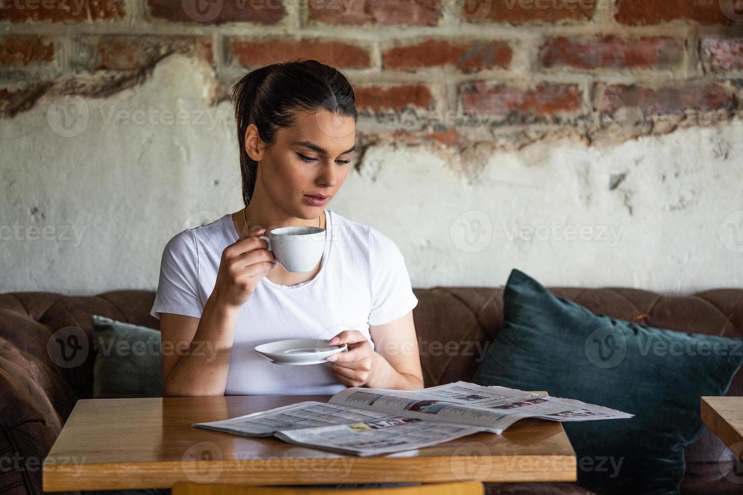Charming woman with beautiful smile reading newspaper during rest in coffee shop, happy Caucasian female reading news while relaxing in cafe during free time photo
