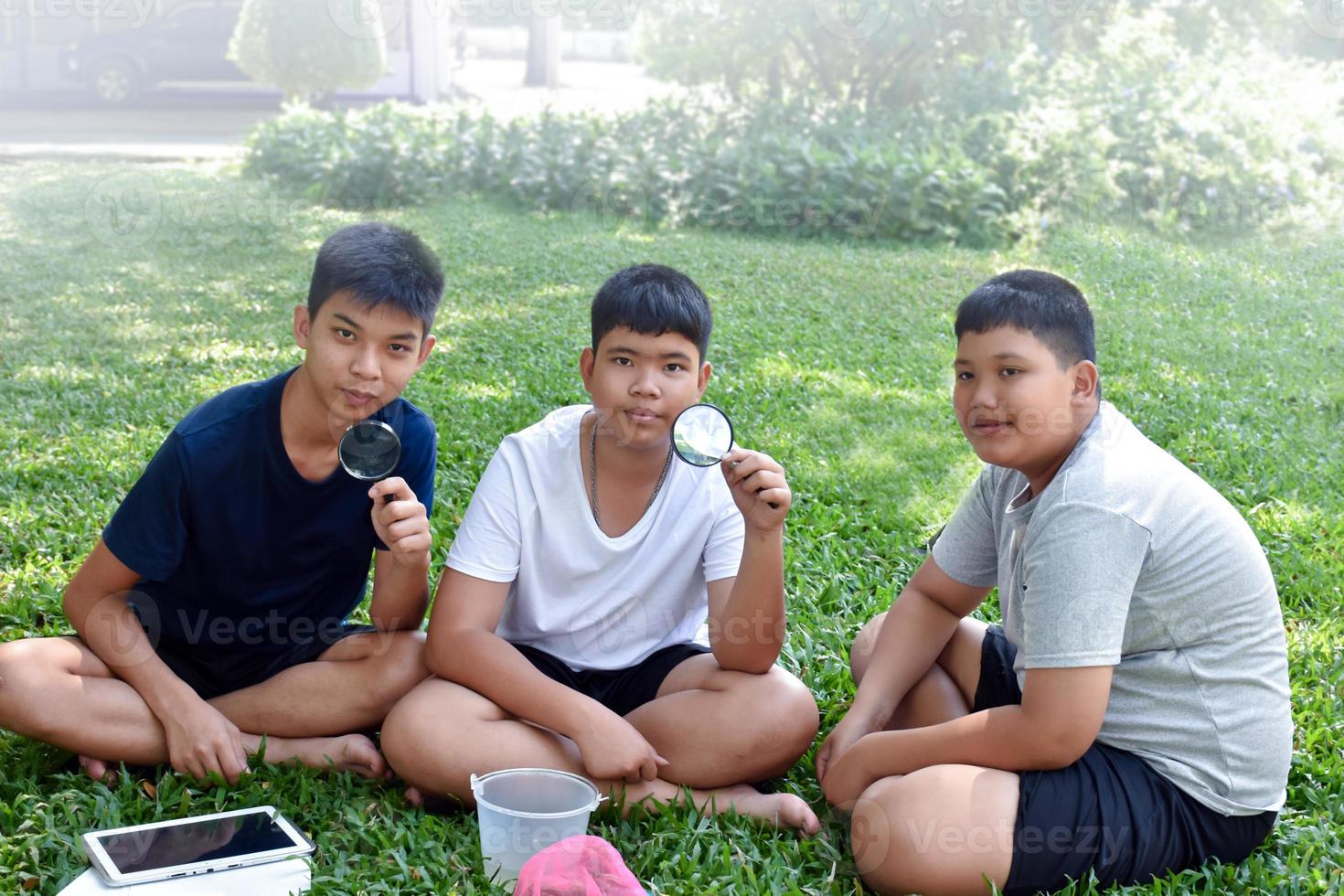 Three Asian studets holding magnifying glasses and other learning devices, tablets, small fishing nets and a small white plastic bin to learn about underwater insects after taking it out of the pond. photo