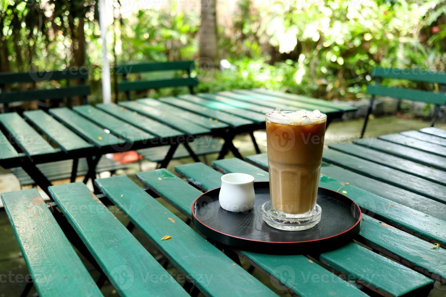 A glass of ice coffee with syrup in white cup on black round plate at green painted wood table in the garden. photo