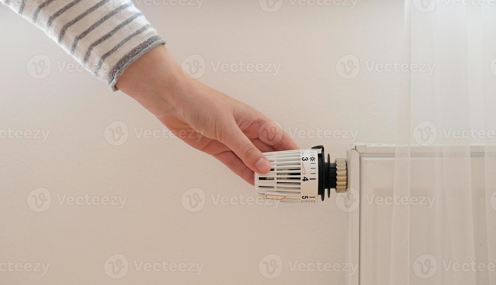 Woman regulates temperature at home with the heating thermostat to save energy, close-up with hand. Temperature control valve on radiator. White heater on wall with curtain in modern apartment. photo