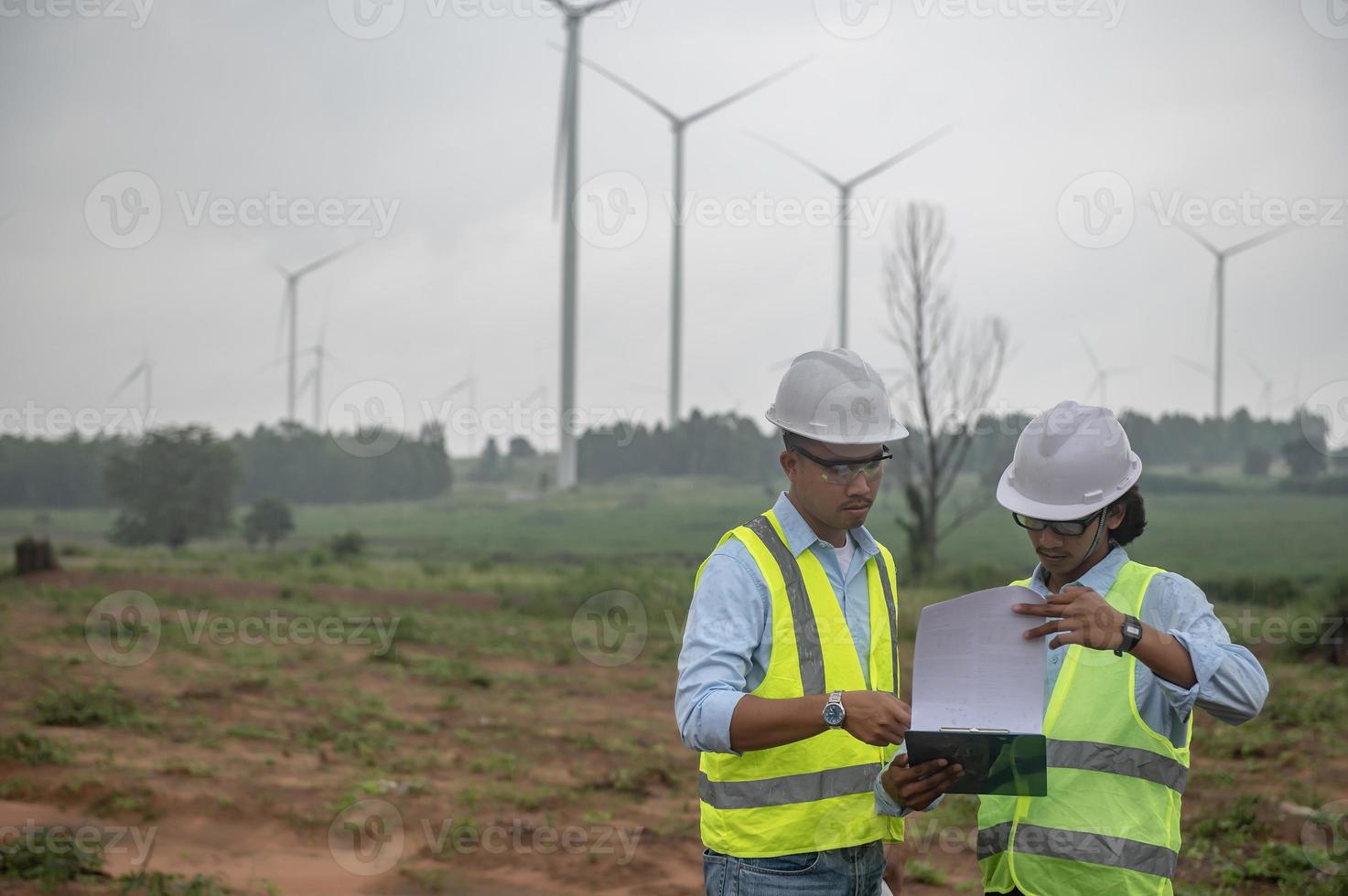 Two engineers working and holding the report at wind turbine farm Power Generator Station on mountain,Thailand people,Technician man and woman discuss about work photo