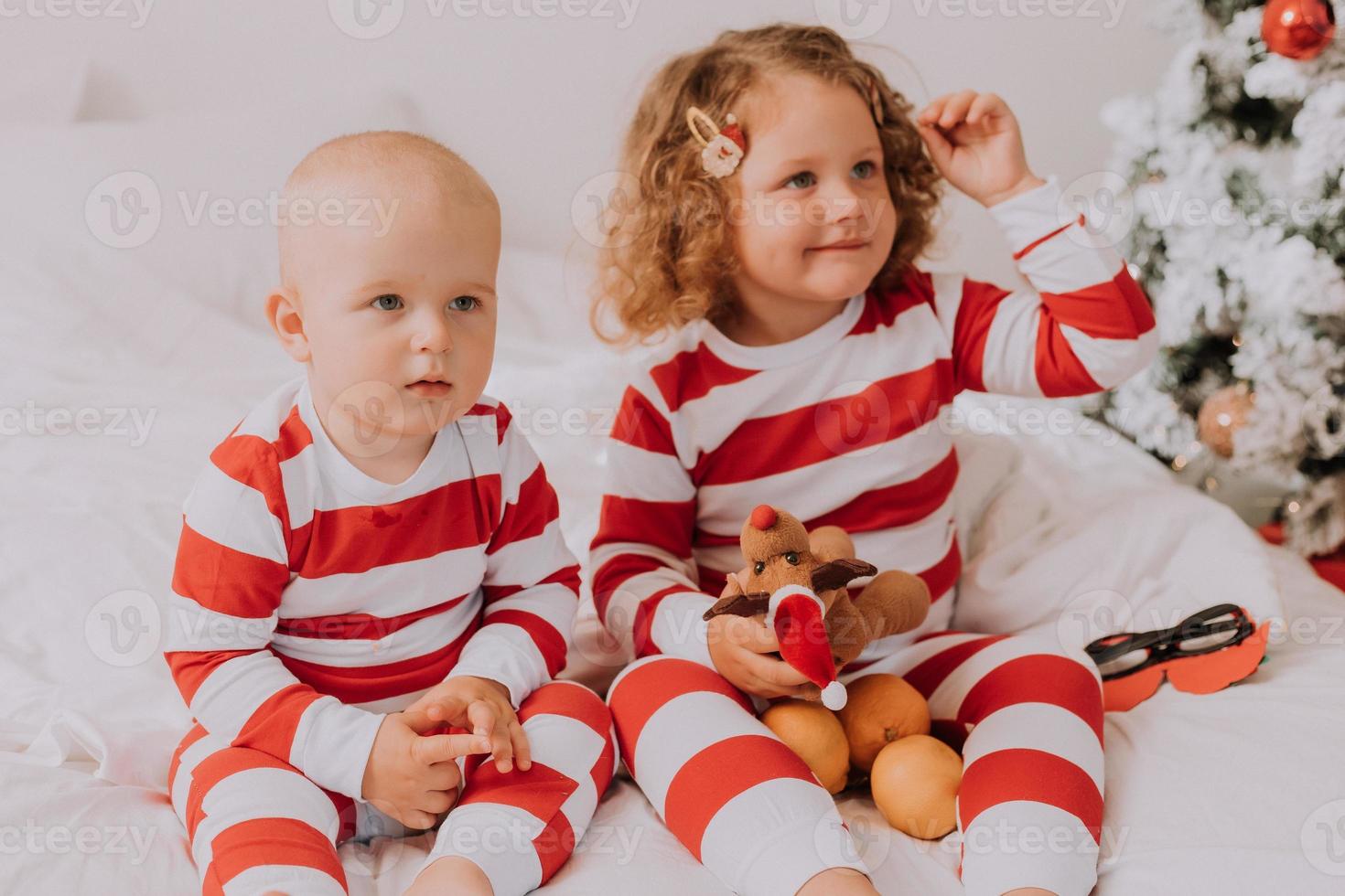 los niños en pijama rojo y blanco prueban gafas divertidas con santa claus sentado en la cama. estilo de vida. hermano y hermana celebrando la navidad. niño y niña están jugando en casa. foto de alta calidad