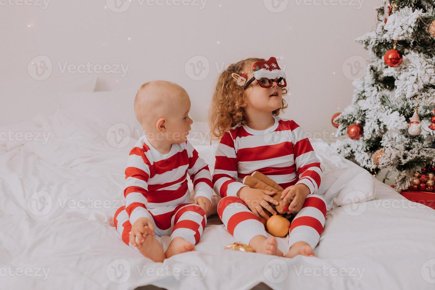 children in red and white pajamas try on funny glasses with Santa Claus sitting in bed. lifestyle. brother and sister celebrating Christmas. boy and girl are playing at home. High quality photo