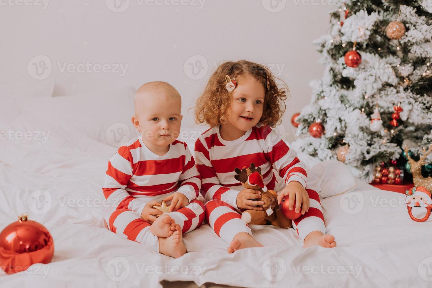 children in red and white pajamas try on funny glasses with Santa Claus sitting in bed. lifestyle. brother and sister celebrating Christmas. boy and girl are playing at home. High quality photo