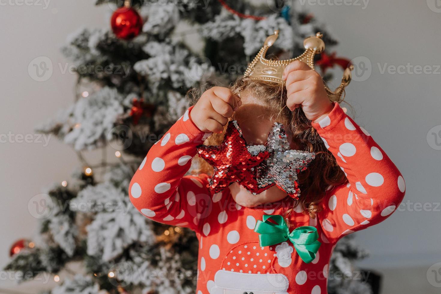 little curly-haired girl in a carnival dress hid her face behind shiny Christmas tree toys stars. child in a red dress with a Santa print on the background of a Christmas tree. High quality photo