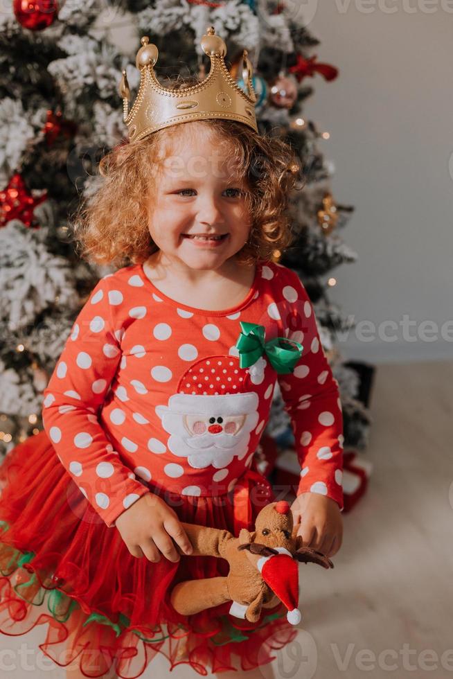 una niña de cabello rizado con un vestido de carnaval escondió su rostro detrás de brillantes estrellas de juguetes de árboles de Navidad. niño con un vestido rojo con un estampado de santa en el fondo de un árbol de navidad. foto de alta calidad