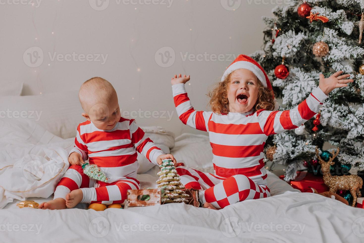 children in red and white pajamas try on funny glasses with Santa Claus sitting in bed. lifestyle. brother and sister celebrating Christmas. boy and girl are playing at home. High quality photo