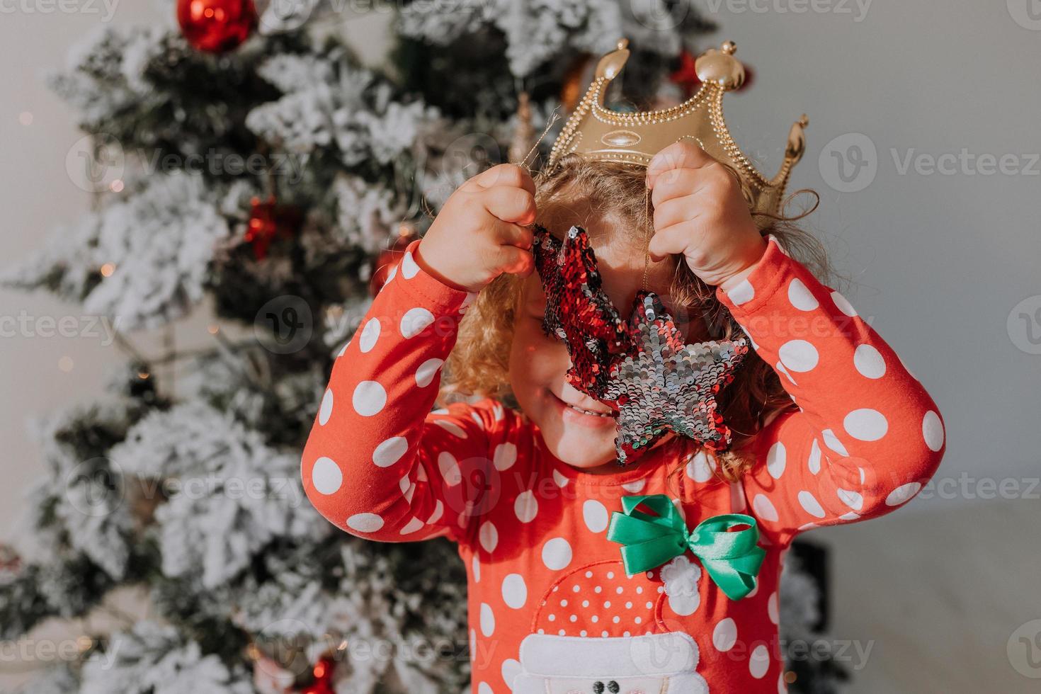 little curly-haired girl in a carnival dress hid her face behind shiny Christmas tree toys stars. child in a red dress with a Santa print on the background of a Christmas tree. High quality photo