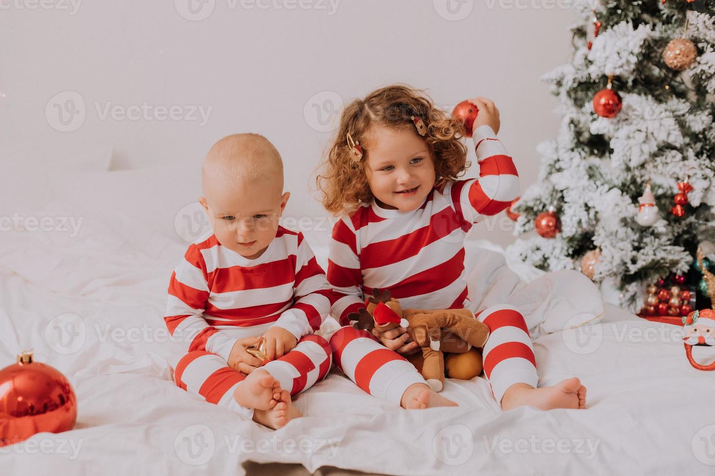 children in red and white pajamas try on funny glasses with Santa Claus sitting in bed. lifestyle. brother and sister celebrating Christmas. boy and girl are playing at home. High quality photo