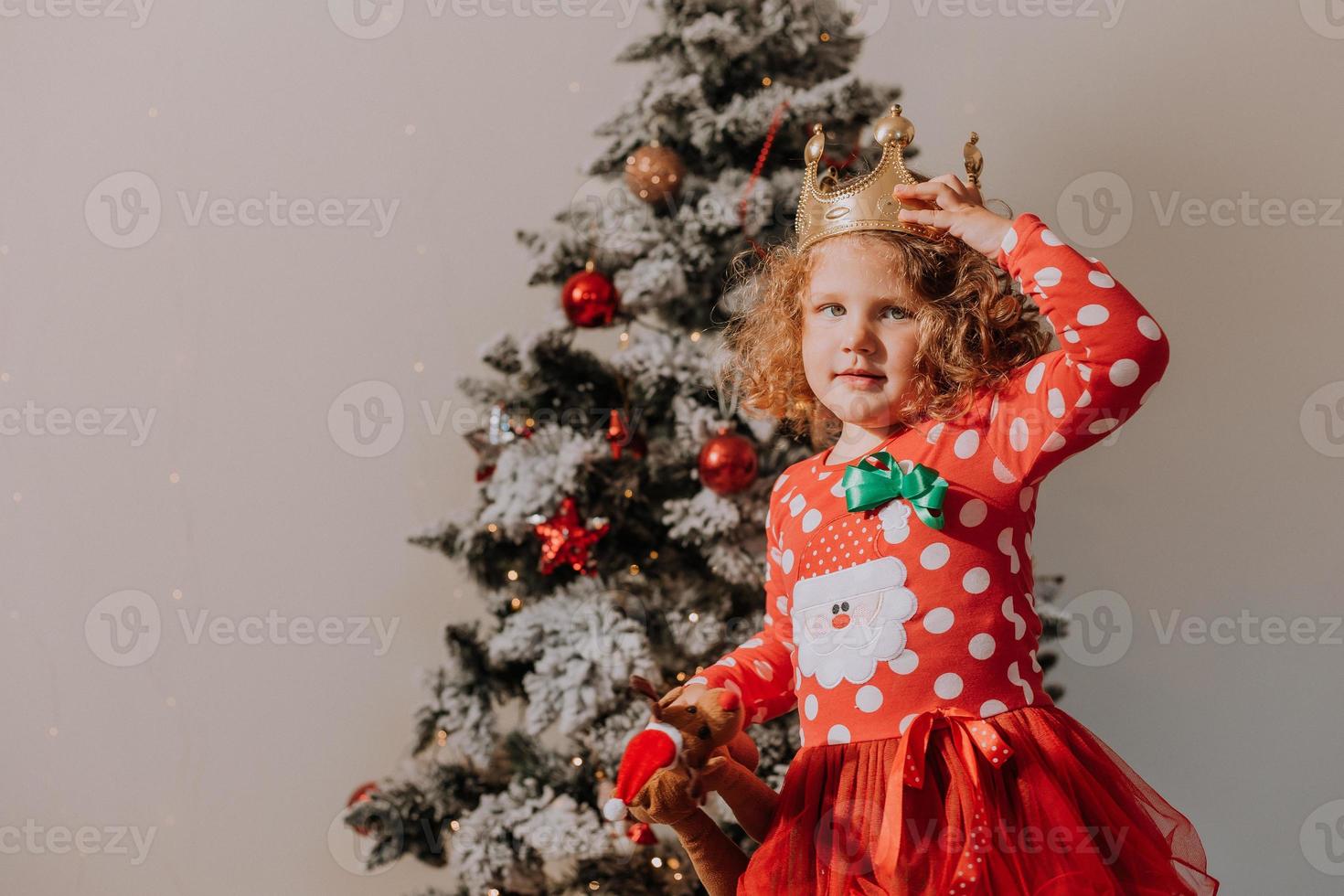 little curly-haired girl in a carnival dress hid her face behind shiny Christmas tree toys stars. child in a red dress with a Santa print on the background of a Christmas tree. High quality photo