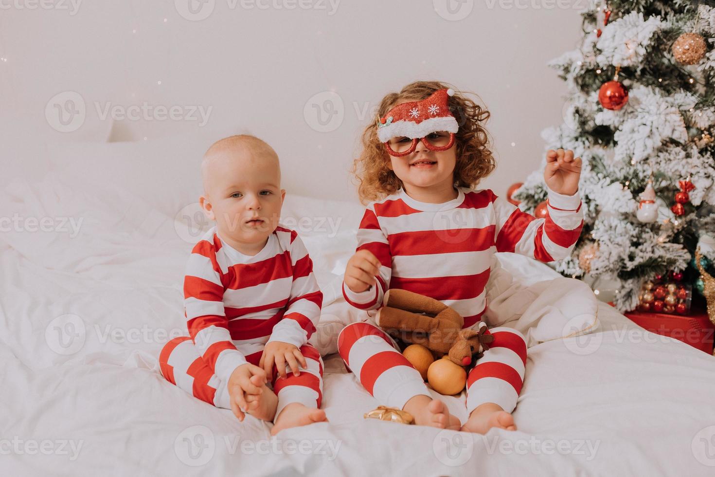 children in red and white pajamas try on funny glasses with Santa Claus sitting in bed. lifestyle. brother and sister celebrating Christmas. boy and girl are playing at home. High quality photo